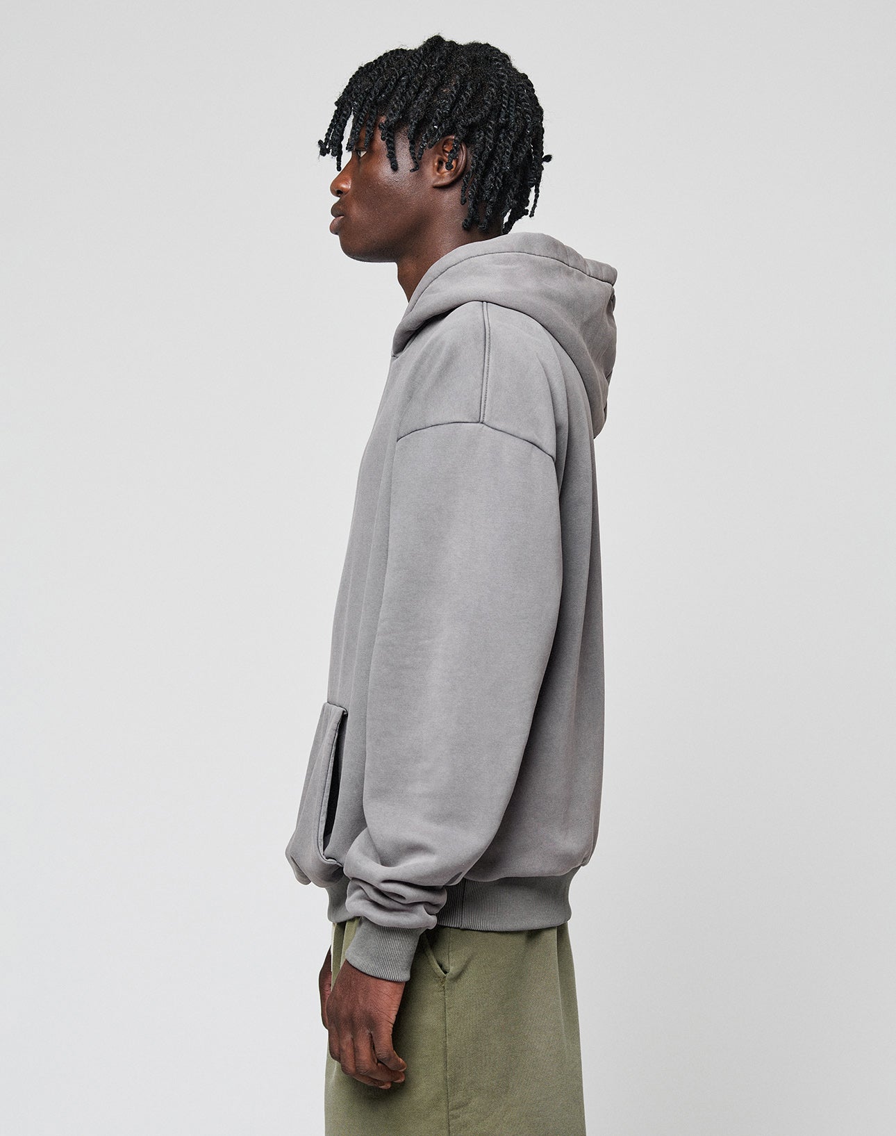 A person with short, twisted hair wears the LIVE FAST DIE YOUNG Basic Blank Hooded in oversize fit and olive green pants, standing against a plain light gray background.| model