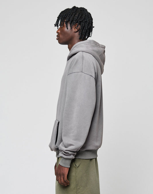 A person with short, textured hair stands sideways in a loose LIVE FAST DIE YOUNG Basic Blank Hooded hoodie and olive green pants against a plain light gray background.