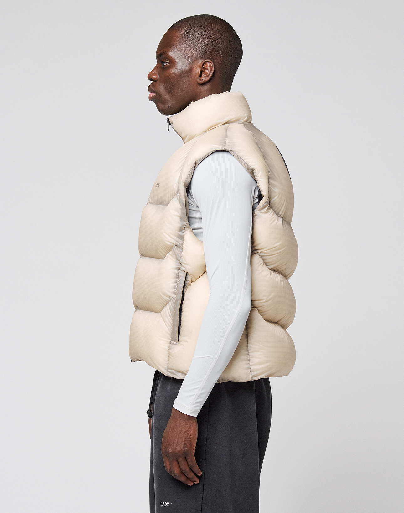 A man stands in profile against a plain background, wearing the LIVE FAST DIE YOUNG Live Fast Puffer Vest in oversize light beige with transparent outer fabric, over a light gray long-sleeve shirt and dark pants.