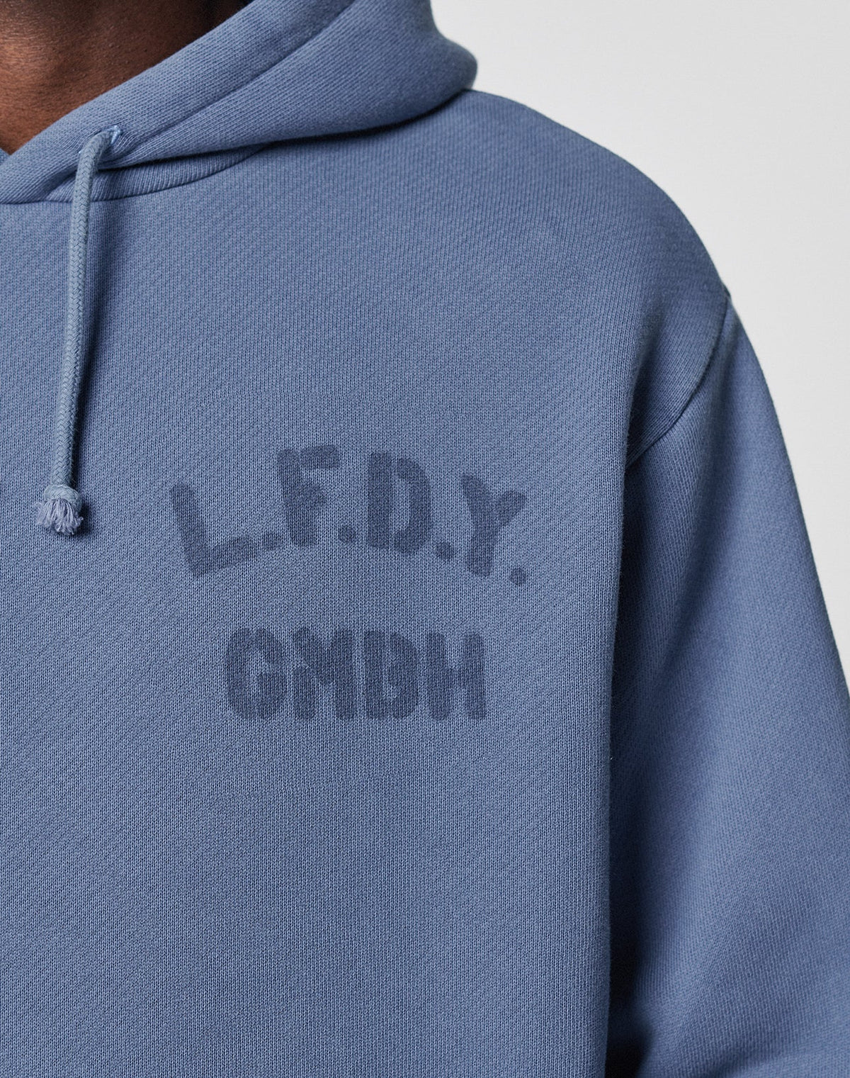A close-up of a person wearing the blue LFDY GmbH Hooded by LIVE FAST DIE YOUNG, featuring a Kapuze mit Kordeln and faded, matching blue L.F.D.Y. GMBH text printed on the chest.