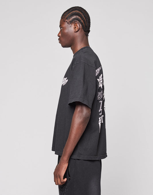 A young man with braided hair stands in profile against a light gray background, wearing a LIVE FAST DIE YOUNG LFDY Logo Tee—an oversized black graphic t-shirt made from 100% Baumwolle—and black shorts.