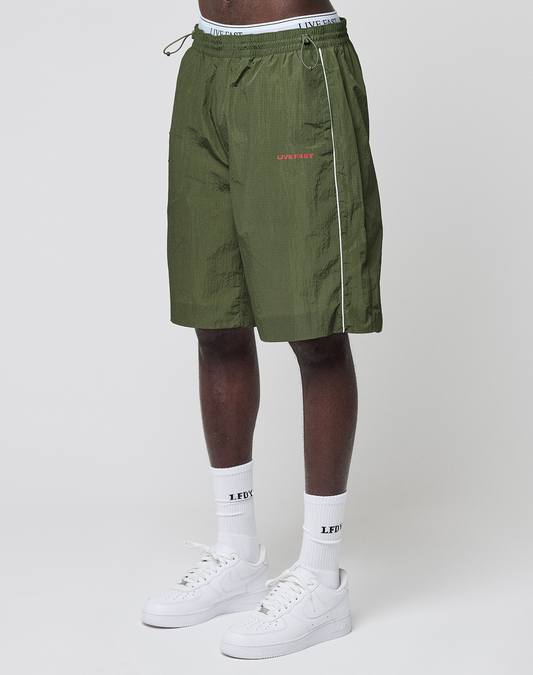 A person wears LIVE FAST DIE YOUNG LFDY Nylon Trackshorts in wide-fit olive green with white piping, a drawstring waist, and red LFDY CLUB logo-print, styled with white socks and sneakers against a plain background. | model