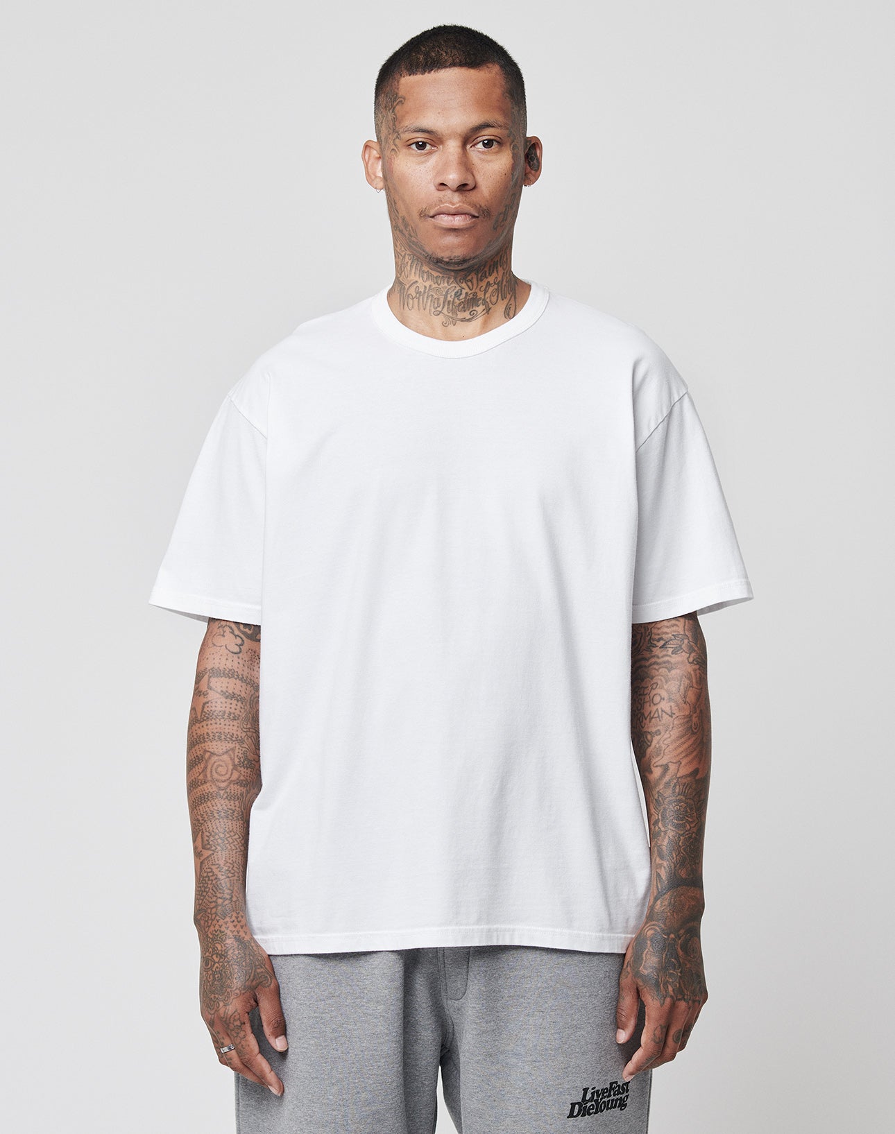 A man with tattoos on his neck, arms, and hands stands facing forward, wearing the LFDY Blank Tee by LIVE FAST DIE YOUNG in white (100% cotton, Regular Fit) and light gray sweatpants against a plain light gray background.