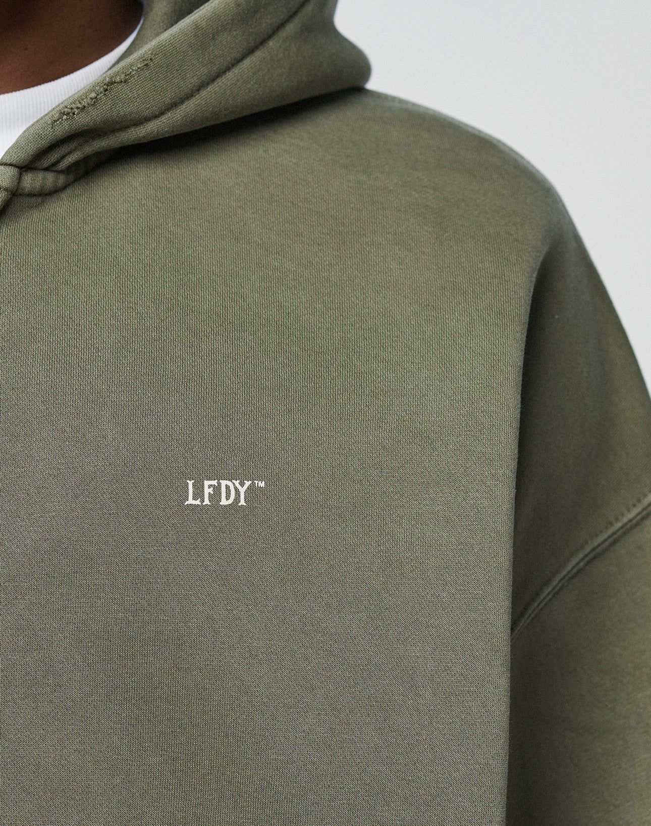 Close-up of a person wearing the olive green LIVE FAST DIE YOUNG Basic Hooded Zip, featuring white LFDY™ embroidery on the chest and a subtly faded fabric for a casual, worn-in look.