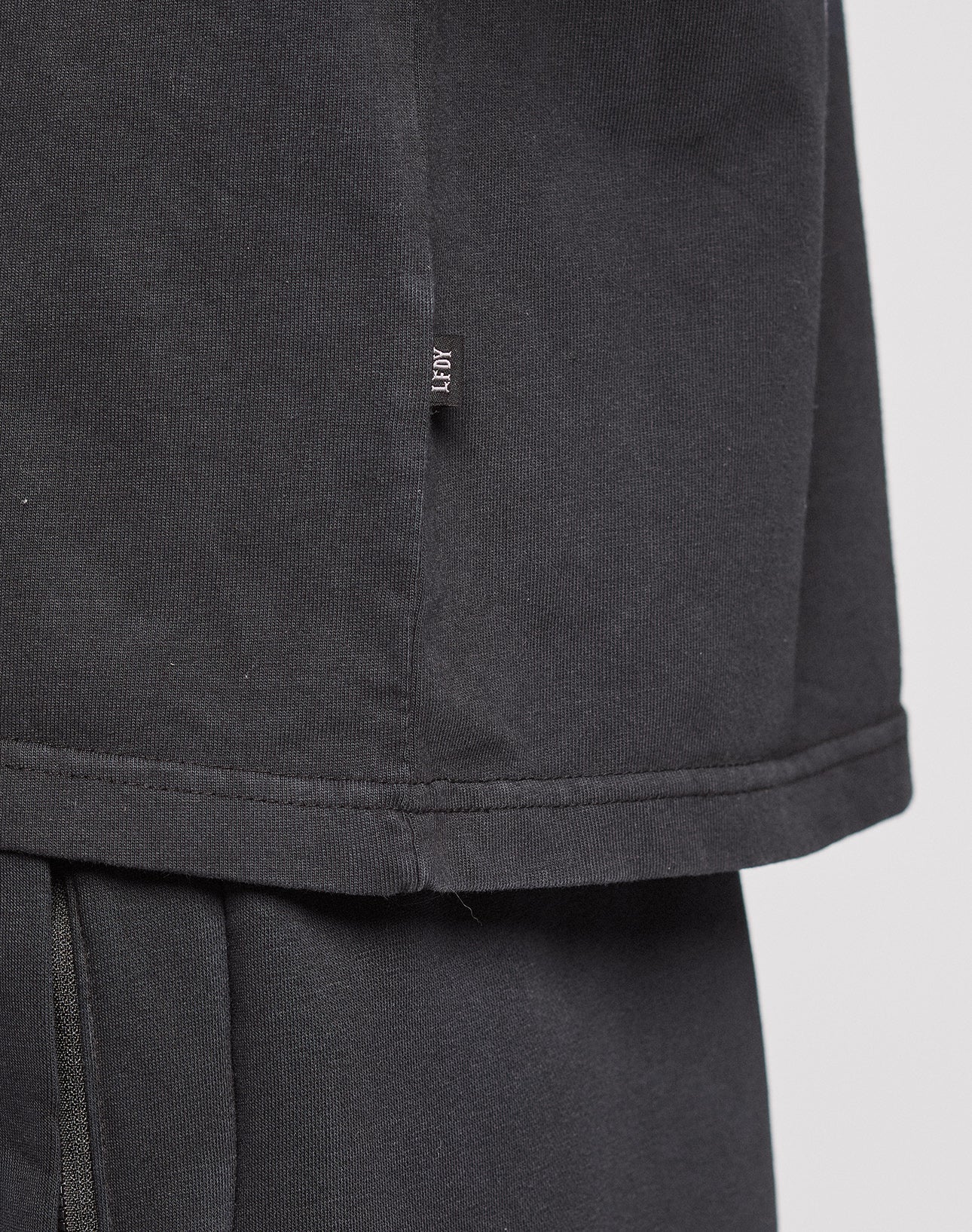 Close-up of the LFDY Logo Tee by LIVE FAST DIE YOUNG in black, featuring an Oversize Fit with a small black fabric tag and white text on the side seam. Layered over matching black pants against a plain light gray background.