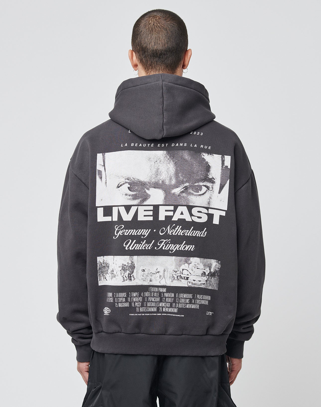 A person with short hair is seen from behind wearing the LIVE FAST DIE YOUNG LFDY Cinema Hooded in Oversize Fit, featuring a bold eye graphic, the words LIVE FAST, country names, and garment details on the back. | model