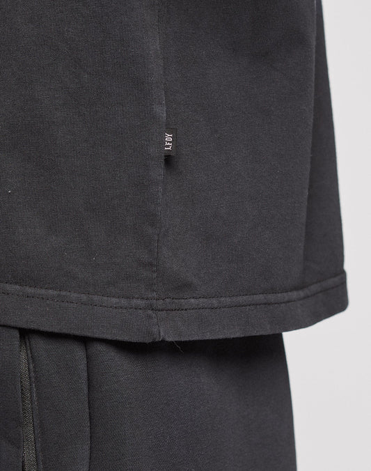 Close-up of the LFDY Logo Tee by LIVE FAST DIE YOUNG in black, featuring an Oversize Fit with a small black fabric tag and white text on the side seam. Layered over matching black pants against a plain light gray background.