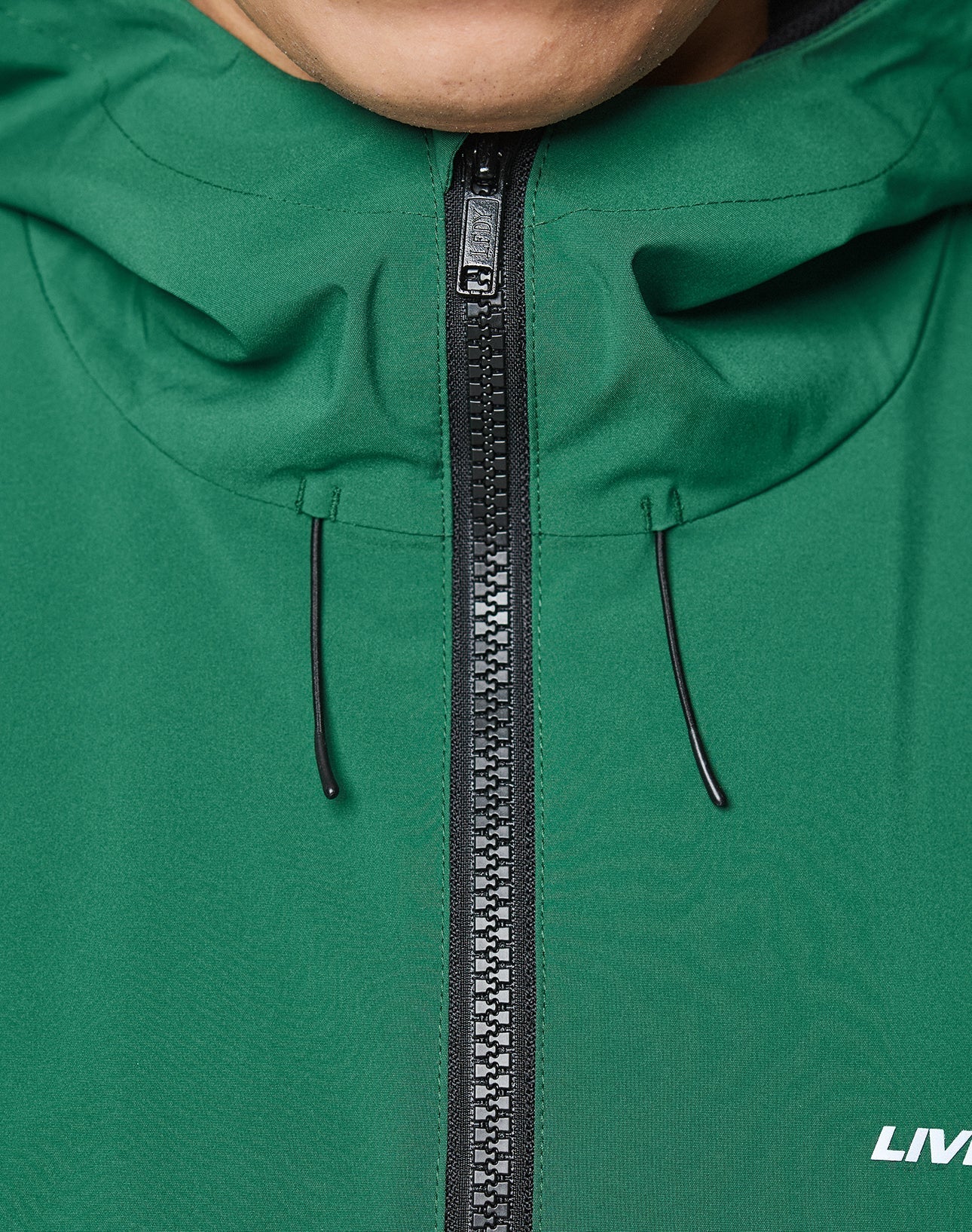 Close-up of the LIVE FAST DIE YOUNG Basic Shell Jacket in green with an oversized fit, front zipper, and black drawstrings. The lower right corner features a white LIV reflective print. The bottom part of a person’s face is seen above the collar.