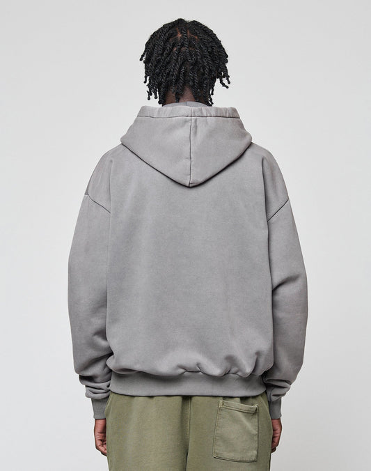 A person with short, twisted hair is seen from behind in a LIVE FAST DIE YOUNG Basic Blank Hooded gray hoodie (oversize cotton-polyester) and olive green back-pocket pants, standing against a plain light background.