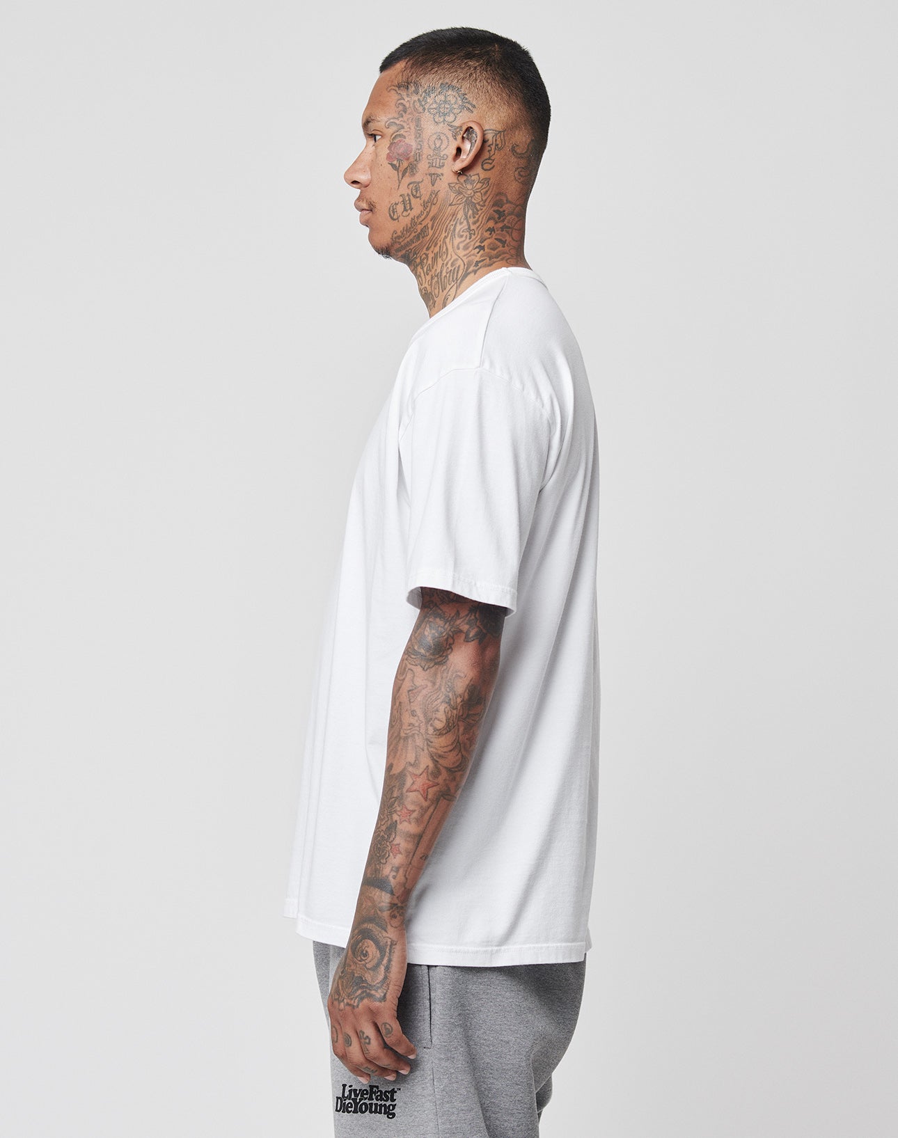 A tattooed man stands in profile, facing left, wearing the LIVE FAST DIE YOUNG LFDY Blank Tee in regular fit (100% cotton) and gray shorts against a light gray background.