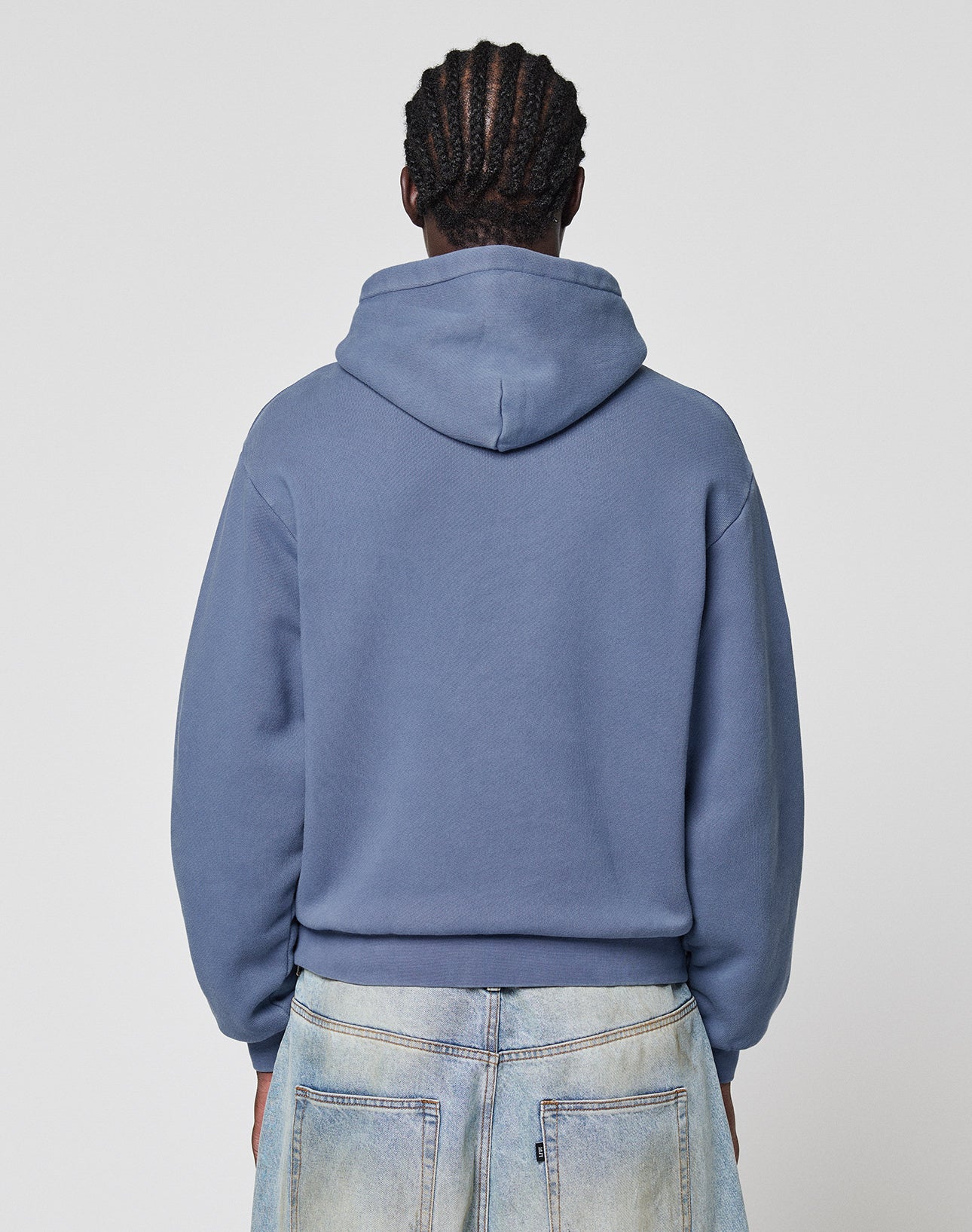 A person with braided hair is seen from the back wearing a blue LIVE FAST DIE YOUNG LFDY GmbH Hooded and light-washed jeans against a plain, light background.