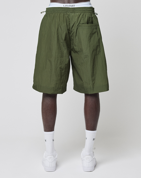 A person stands facing away, wearing LIVE FAST DIE YOUNG LFDY Nylon Trackshorts in olive green, white crew socks, and white sneakers. The shorts feature a back pocket with the logo-print waistband of their white underwear visible above.