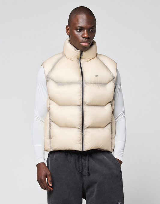 A man faces forward wearing the LIVE FAST DIE YOUNG Live Fast Puffer Vest in cream over a fitted white long-sleeve shirt and dark gray sweatpants, set against a plain light background.
