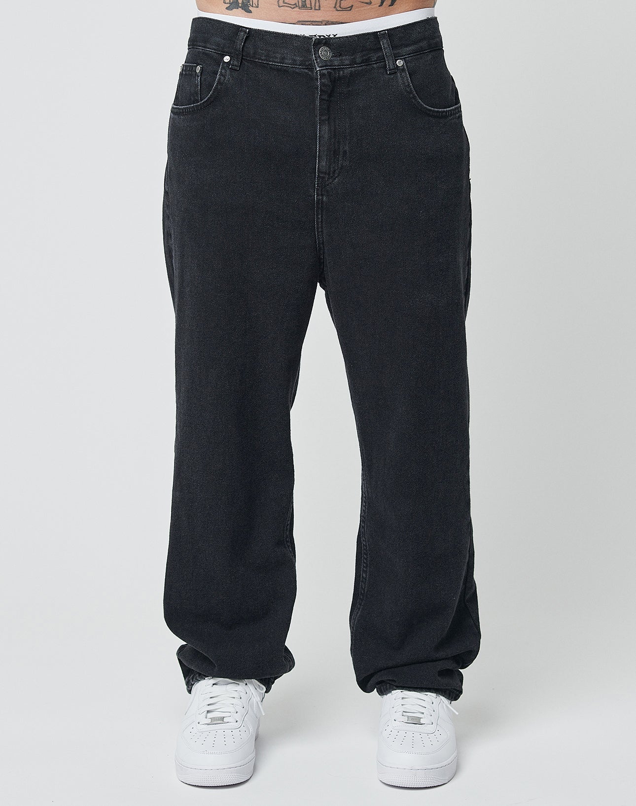 Eine Person trägt LIVE FAST DIE YOUNG Wide Denim in Schwarz und weiße Sneakers vor schlicht weißem Hintergrund; nur die untere Körperhälfte ist zu sehen.