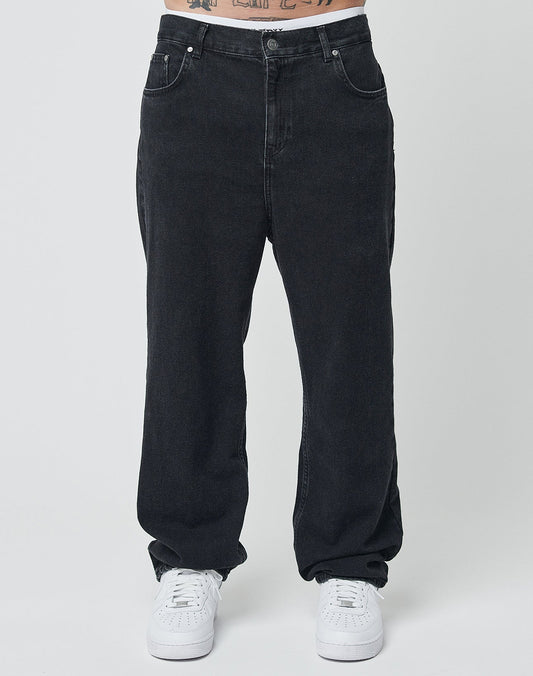 Eine Person trägt LIVE FAST DIE YOUNG Wide Denim in Schwarz und weiße Sneakers vor schlicht weißem Hintergrund; nur die untere Körperhälfte ist zu sehen.