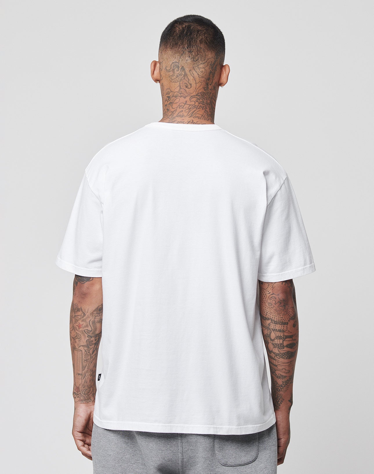 A person with tattooed arms and neck stands with their back to the camera, wearing a LIVE FAST DIE YOUNG LFDY Blank Tee in plain white (100% Baumwolle) and light gray pants against a plain, light gray background.