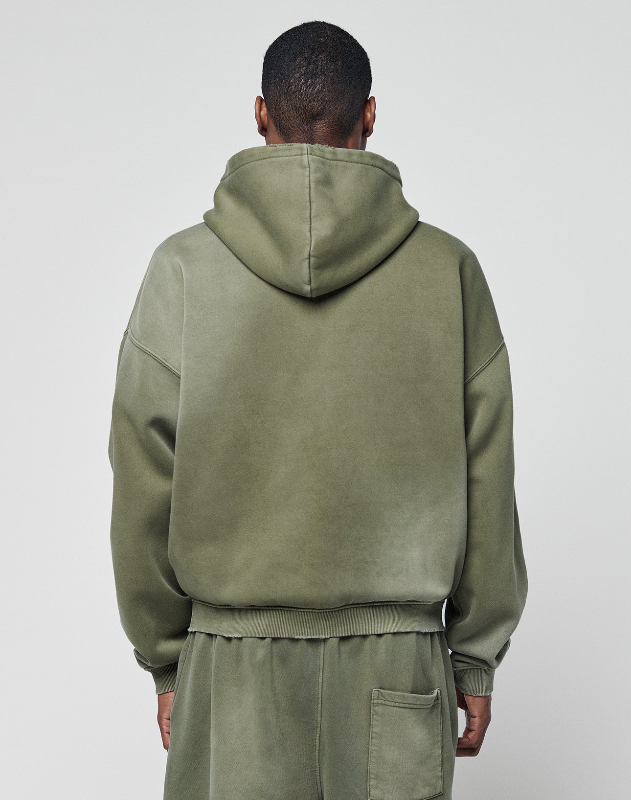 A person faces away from the camera, wearing the LIVE FAST DIE YOUNG Basic Hooded Zip in olive green with a subtle faded effect and matching sweatpants, set against a plain light gray background.