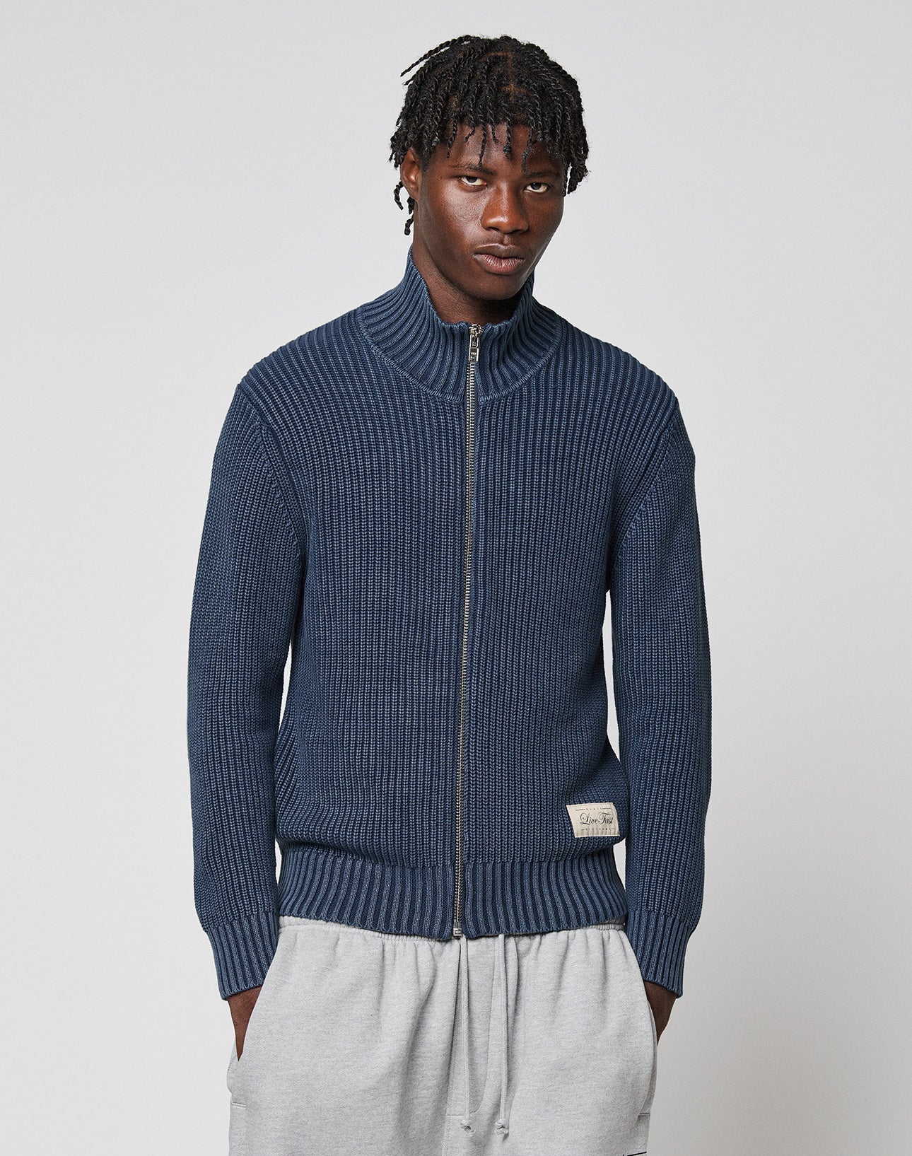 A man with short dreadlocks wears the LFDY Knit Jacket by LIVE FAST DIE YOUNG—a dark blue zip-up ribbed sweater with Stehkragen—and light gray sweatpants, standing against a plain light background.