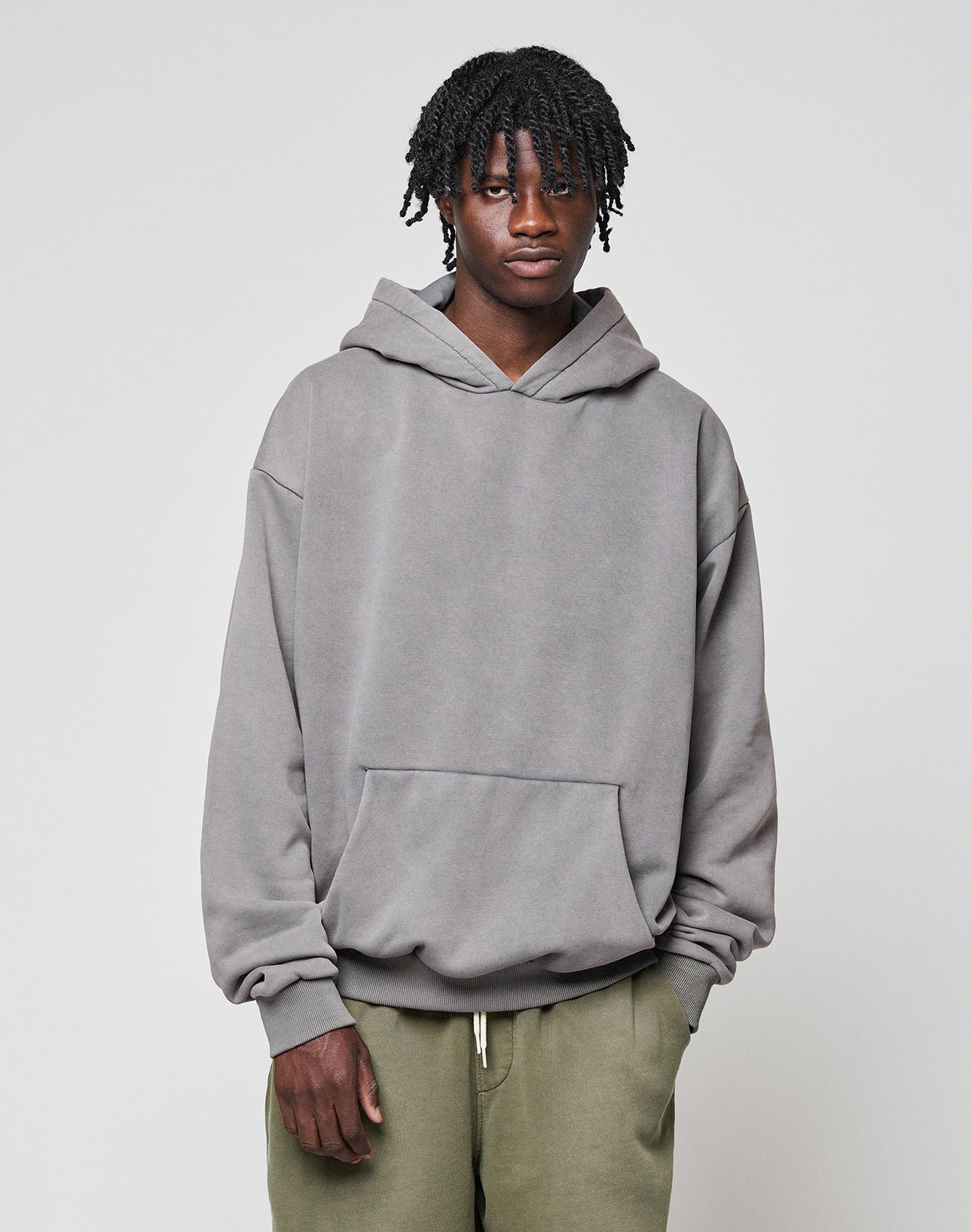A person with short, textured hair stands sideways in a loose LIVE FAST DIE YOUNG Basic Blank Hooded hoodie and olive green pants against a plain light gray background.