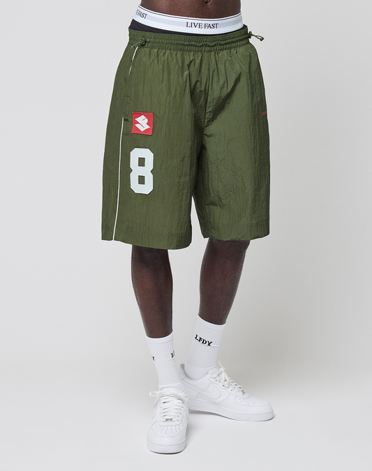 Someone wears LIVE FAST DIE YOUNG LFDY Nylon Trackshorts in wide fit olive green with a white number 8 and red logo-print on the left leg, white Live Fast waistband underwear, white LFDY text socks, and white sneakers against a plain white background.