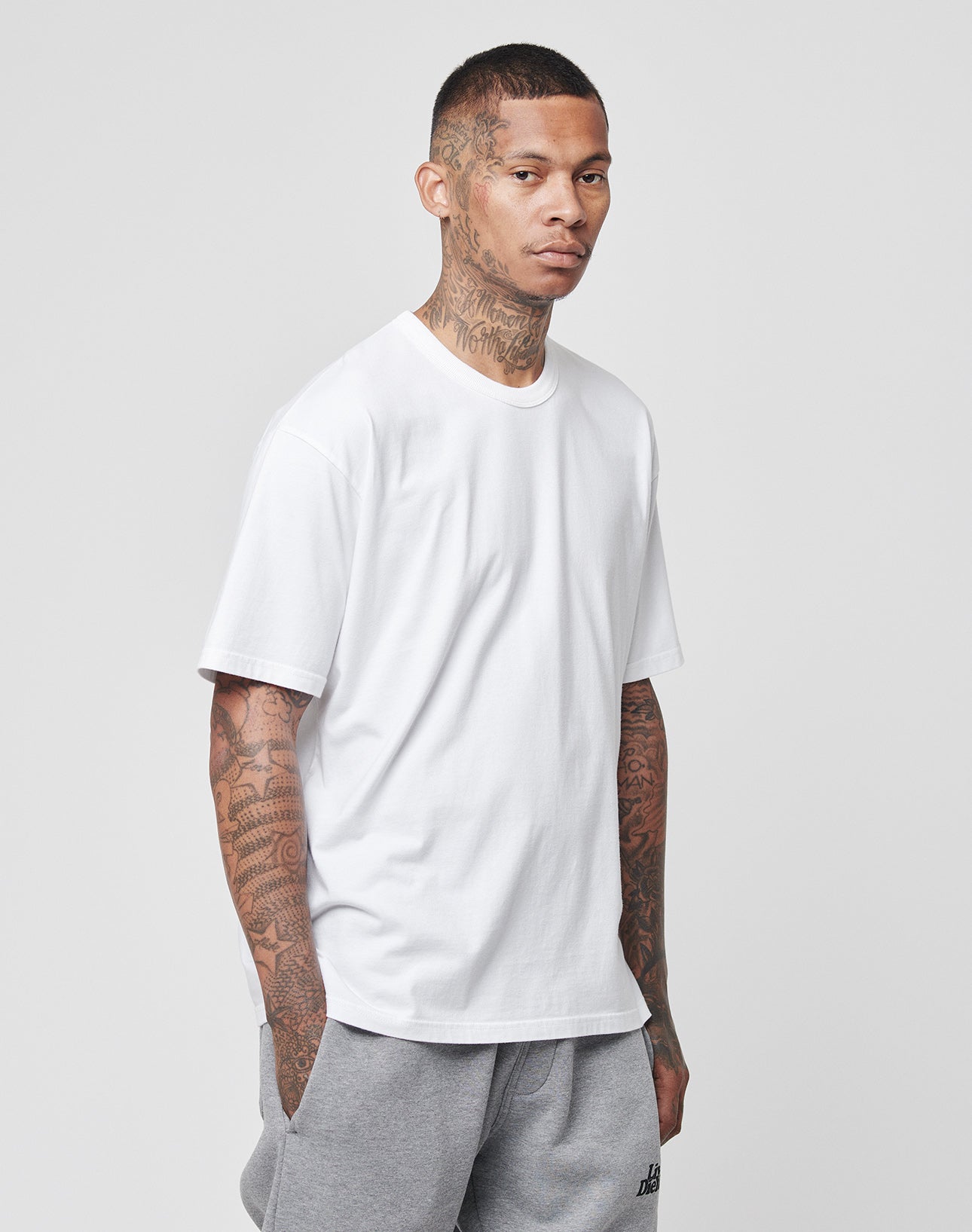A tattooed man stands against a plain gray background, wearing the LIVE FAST DIE YOUNG LFDY Blank Tee in white Regular Fit and light gray sweatpants, with his hands in his pockets.