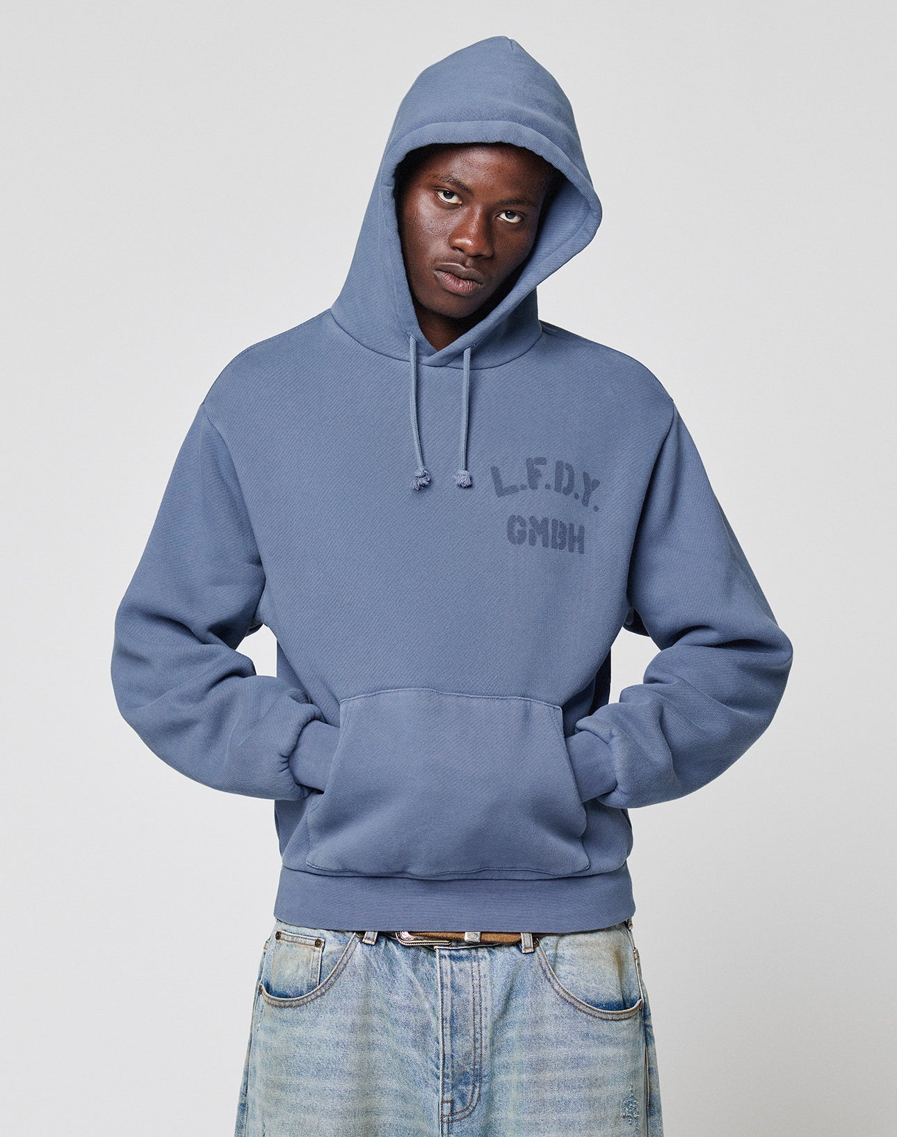 A person wearing a LIVE FAST DIE YOUNG LFDY GmbH Hooded with drawstring hood and front logo stands with hands in the front pocket, looking at the camera. They also wear light blue jeans.