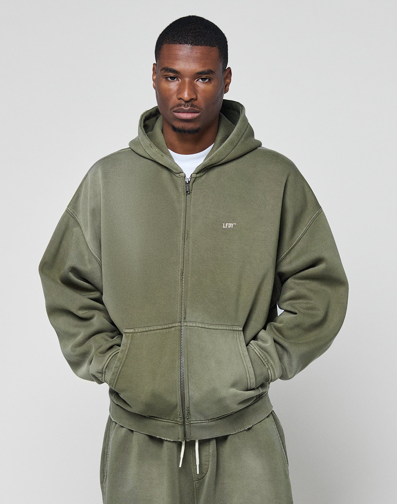 A man in a LIVE FAST DIE YOUNG Basic Hooded Zip hoodie in olive green, with matching sweatpants and a white t-shirt, stands against a light gray background, hands in his hoodie pockets.