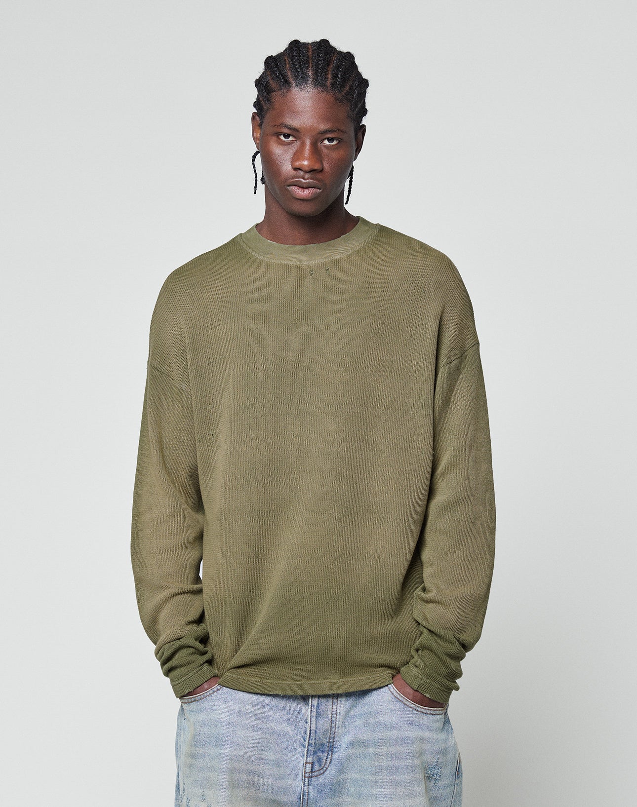 A man with cornrow braids wears the LFDY Rib Knit Longsleeve by LIVE FAST DIE YOUNG in olive green and light blue jeans, standing against a plain light grey background with his hands in his pockets.
