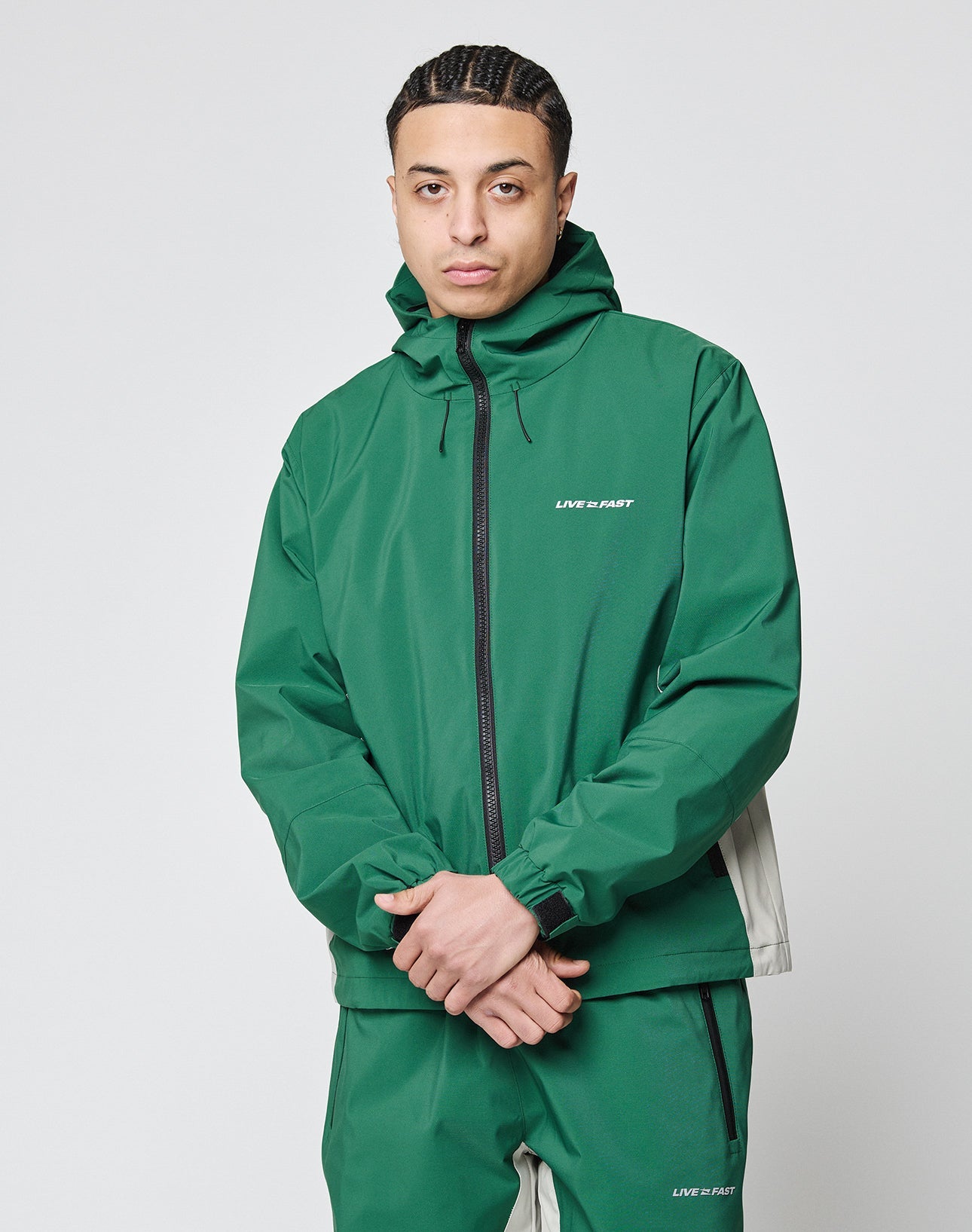 A person with braided hair wears an oversize Basic Shell Jacket and matching pants by LIVE FAST DIE YOUNG, both in green with reflektierender Druck and the words LIVE FAST, standing against a plain white background.