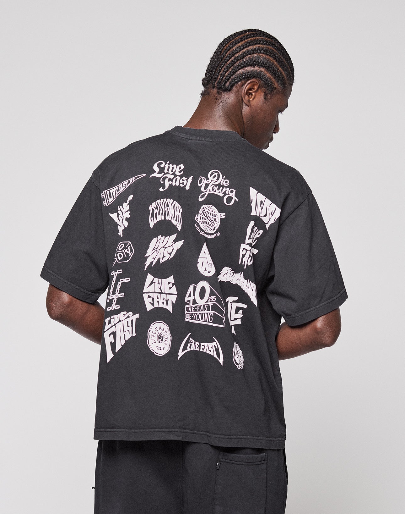 A person with short braids faces away, wearing the LIVE FAST DIE YOUNG LFDY Logo Tee—an oversize black T-shirt with bold white graphics and “Live Fast” text on the back. The background is plain gray.