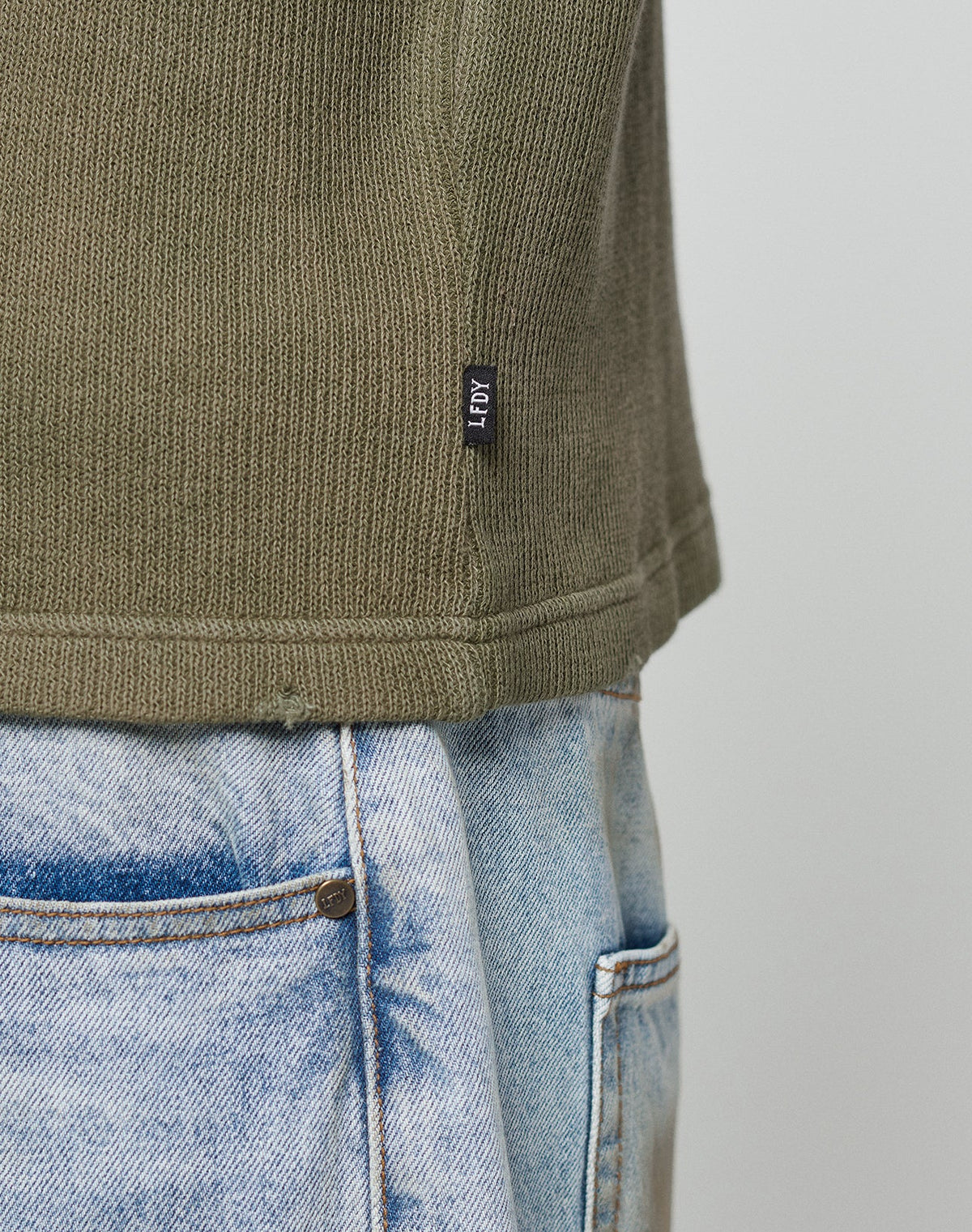Close-up of the side of a person wearing light blue jeans and an olive green LFDY Rib Knit Longsleeve by LIVE FAST DIE YOUNG, featuring an oversized fit and a small black ADY tag near the hem against a plain, light background.