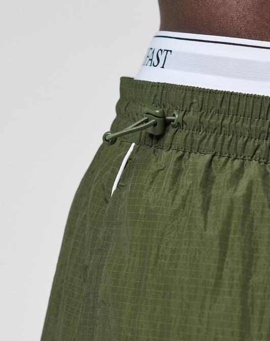 Close-up of a person wearing LFDY Nylon Trackshorts by LIVE FAST DIE YOUNG in olive green, showing a wide fit, adjustable drawstring, textured polyamide fabric, and a white logo-print waistband with black text.