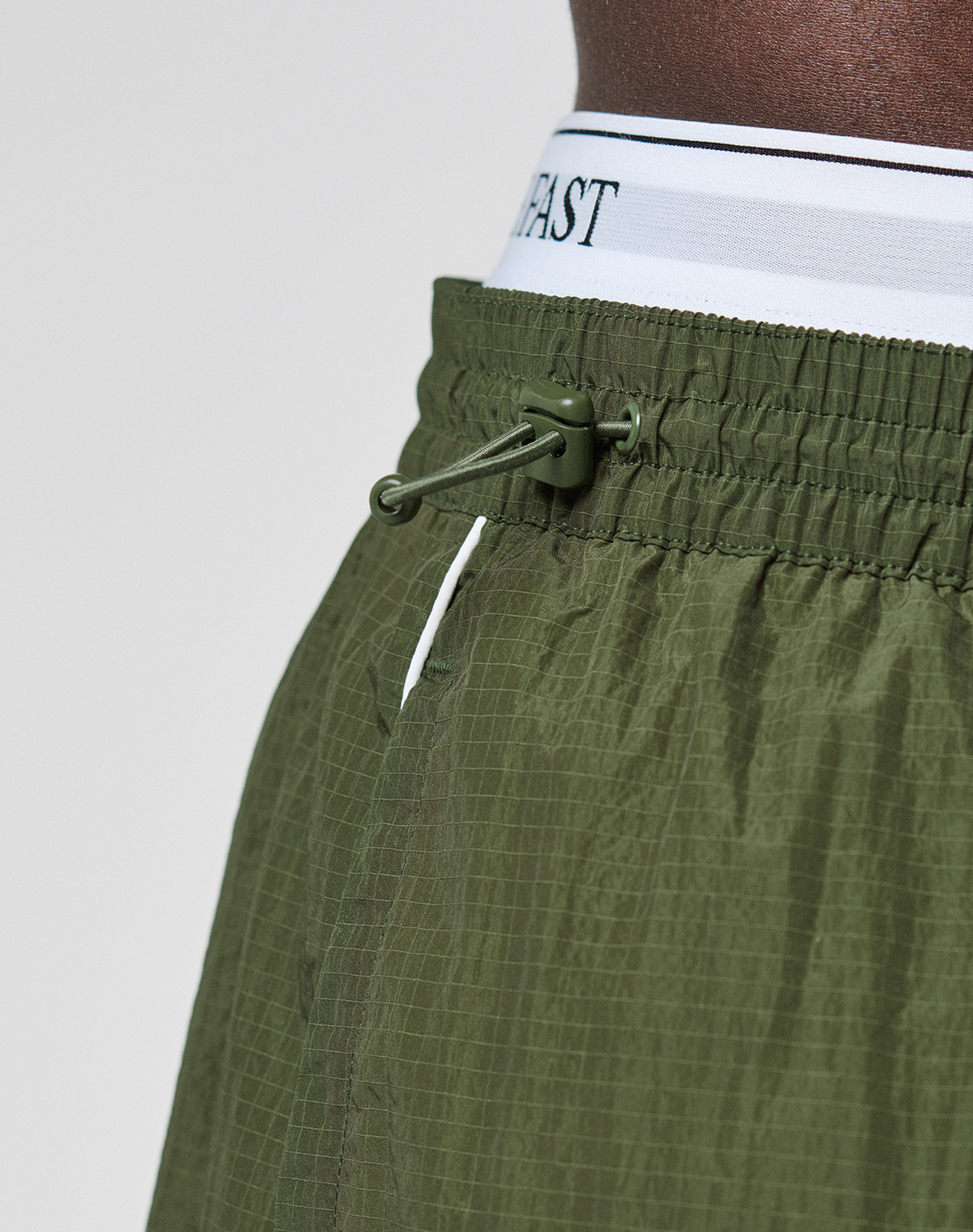 Close-up of a person wearing LFDY Nylon Trackshorts by LIVE FAST DIE YOUNG in olive green, showing a wide fit, adjustable drawstring, textured polyamide fabric, and a white logo-print waistband with black text.