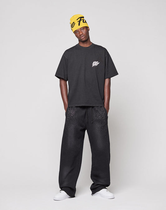 A man stands against a plain background wearing a loose, black LFDY Logo Tee by LIVE FAST DIE YOUNG, black speckled sweatpants, white sneakers, and a yellow beanie with black text.