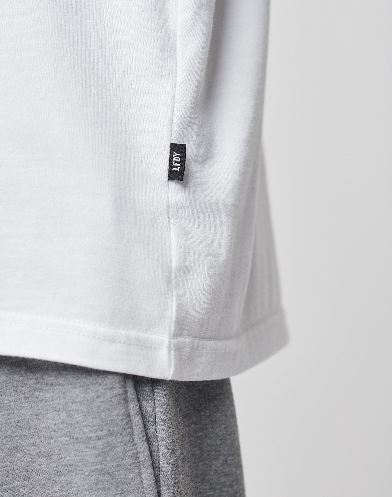 Close-up of a white LFDY Blank Tee by LIVE FAST DIE YOUNG, featuring a small black LFOT tag near the hem, styled with light gray sweatpants. The background is plain and light-colored.