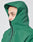 A person wears the LIVE FAST DIE YOUNG Basic Shell Jacket in green with an oversize fit, zipped to the chin with a wasserabweisender Reißverschluss. The hood covers their head and the plain background is light-colored.