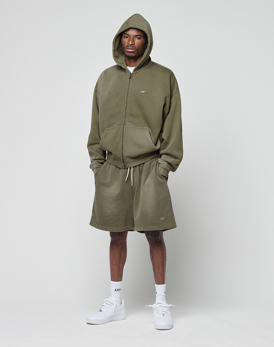 A man stands against a white background wearing the LIVE FAST DIE YOUNG Basic Hooded Zip in olive green with a subtle logo and oversize fit, matching shorts, white socks, and sneakers. Hands in pockets, he looks directly at the camera.