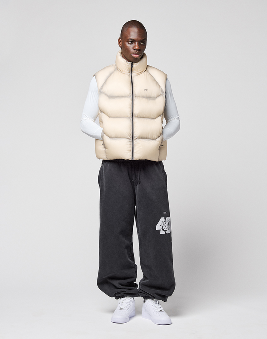 A man faces forward against a plain background, wearing the LIVE FAST DIE YOUNG Live Fast Puffer Vest in oversize light beige with transparent outer layer, over a long-sleeve white shirt, dark gray sweatpants with “46,” and white sneakers.