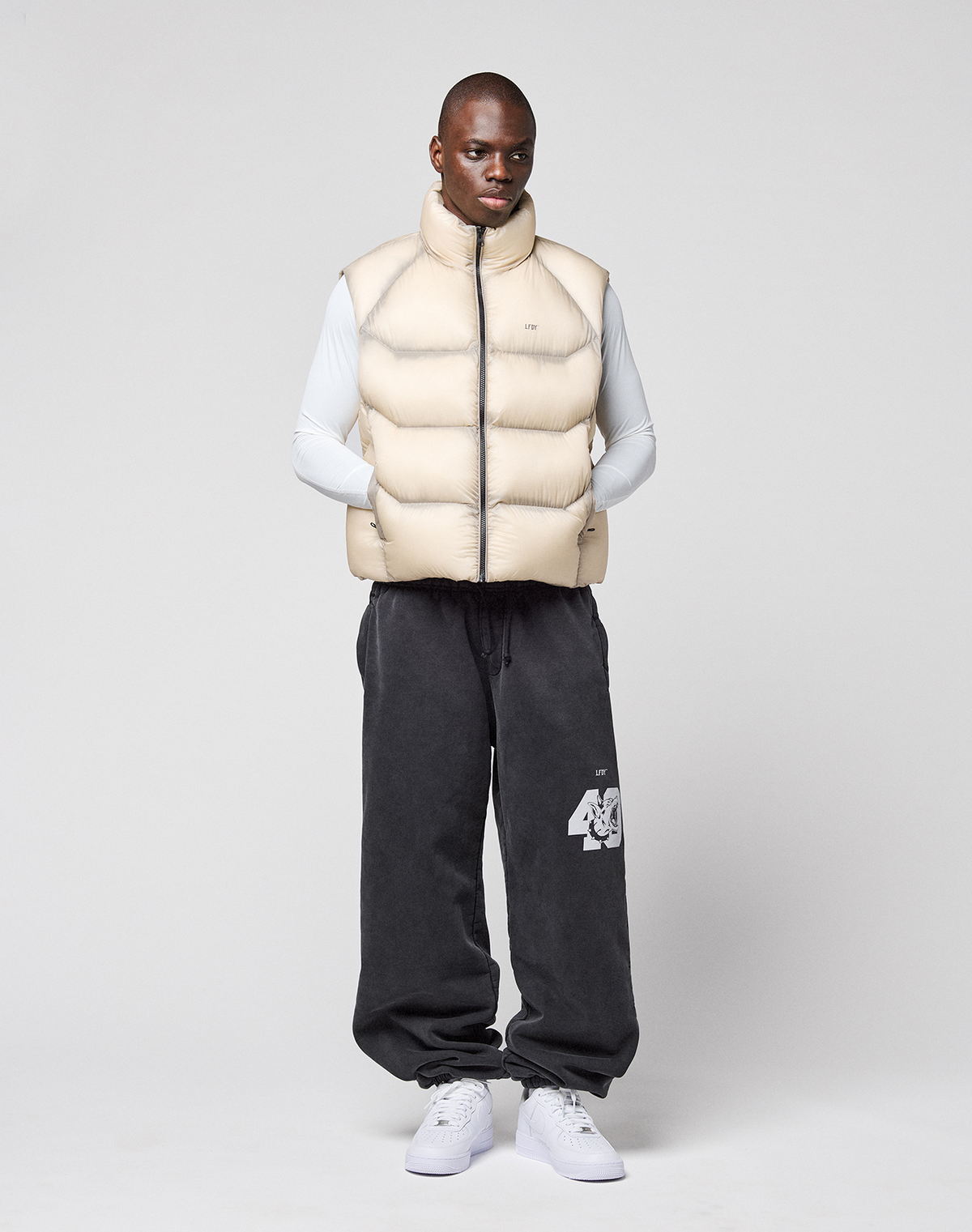 A man faces forward against a plain background, wearing the LIVE FAST DIE YOUNG Live Fast Puffer Vest in oversize light beige with transparent outer layer, over a long-sleeve white shirt, dark gray sweatpants with “46,” and white sneakers.