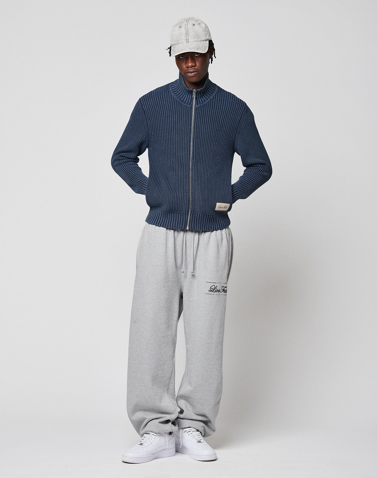 A person in a light gray cap, LIVE FAST DIE YOUNG LFDY Knit Jacket (blue, ribbed, Stehkragen), gray Regular Fit sweatpants with left thigh text, and white sneakers stands against a plain background, hands in pockets, facing the camera.