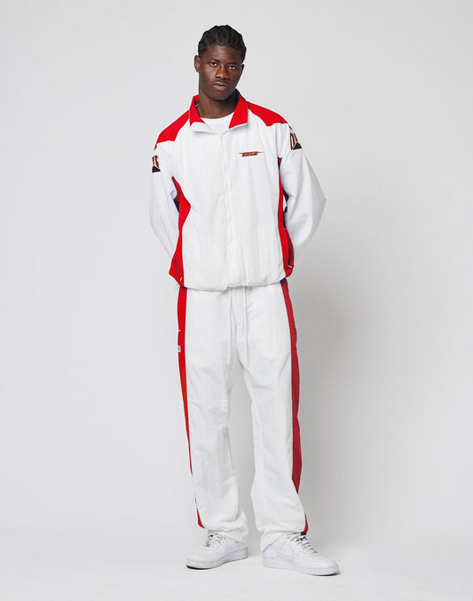 A man with braided hair and white sneakers stands against a plain background, wearing LIVE FAST DIE YOUNG’s LFDY Trackpants 3000—white with red panels, made from Polyamid, featuring an elastic waistband and a wide fit.