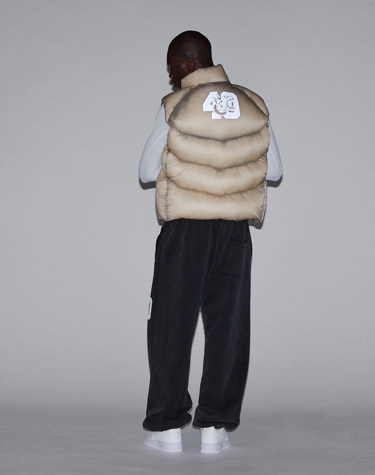 A person faces away, wearing the LIVE FAST DIE YOUNG "Live Fast Puffer Vest" in beige with a white patch, paired with dark pants and white sneakers, set against a plain gray background.