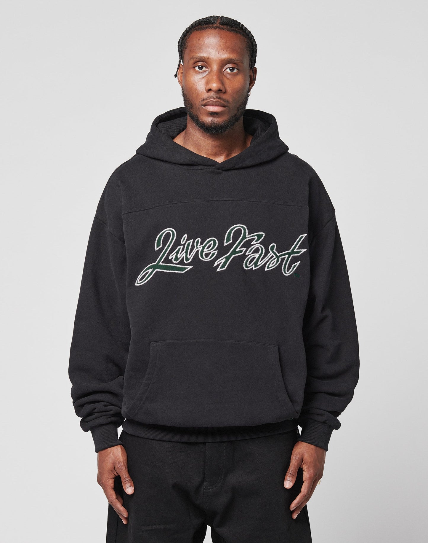 A man with braided hair faces forward in a plain light gray setting, wearing the LIVE FAST DIE YOUNG LFDY Script Hooded. The black 100% Baumwolle hoodie has Distress Effekte, an Oversize Fit, and Live Fast in script on the front.