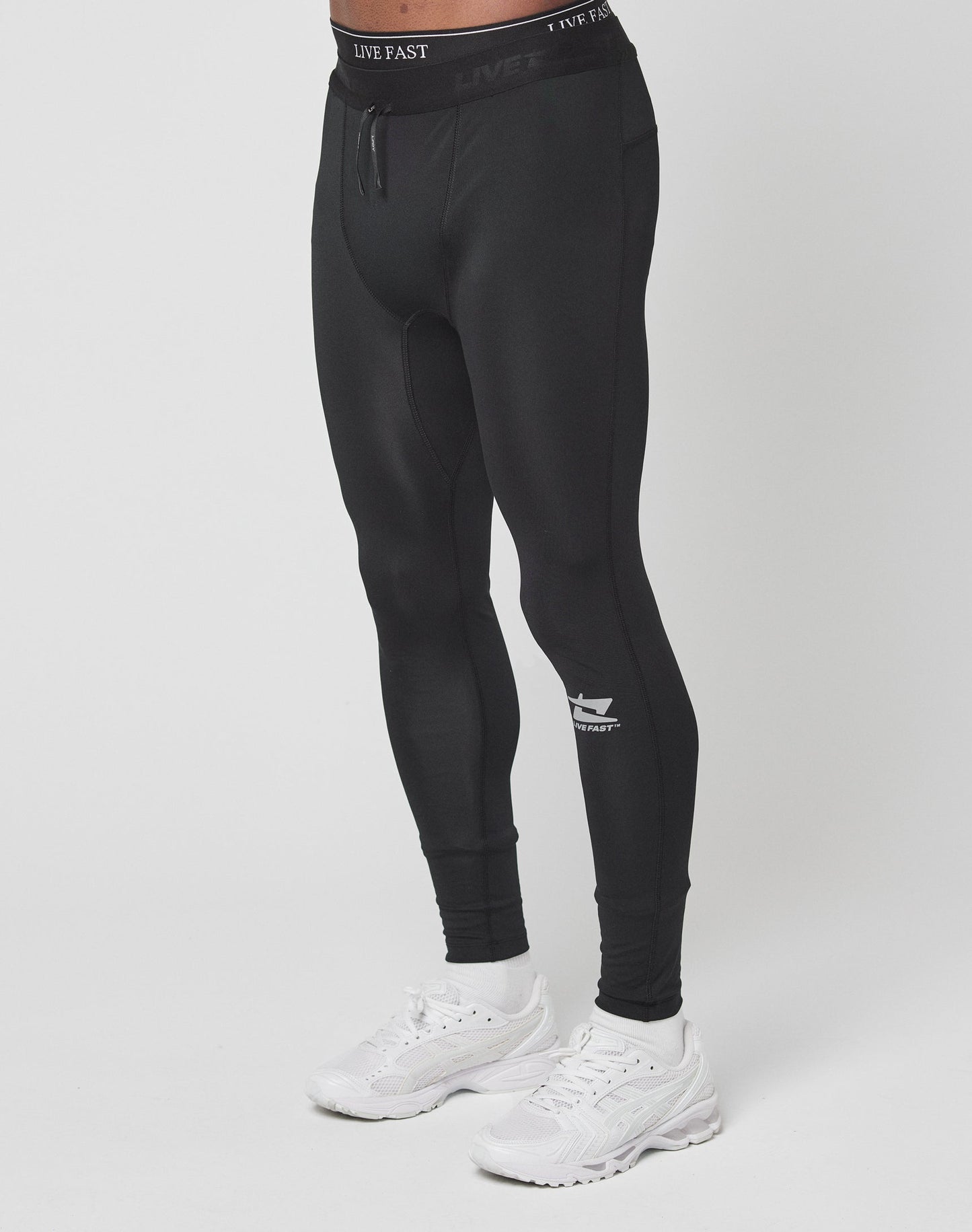 A person wearing Live Fast Performance Tights by LIVE FAST DIE YOUNG, in black slim fit with branding and elastic waistband, paired with white sneakers, stands against a plain white background. Their upper body is not visible.| model