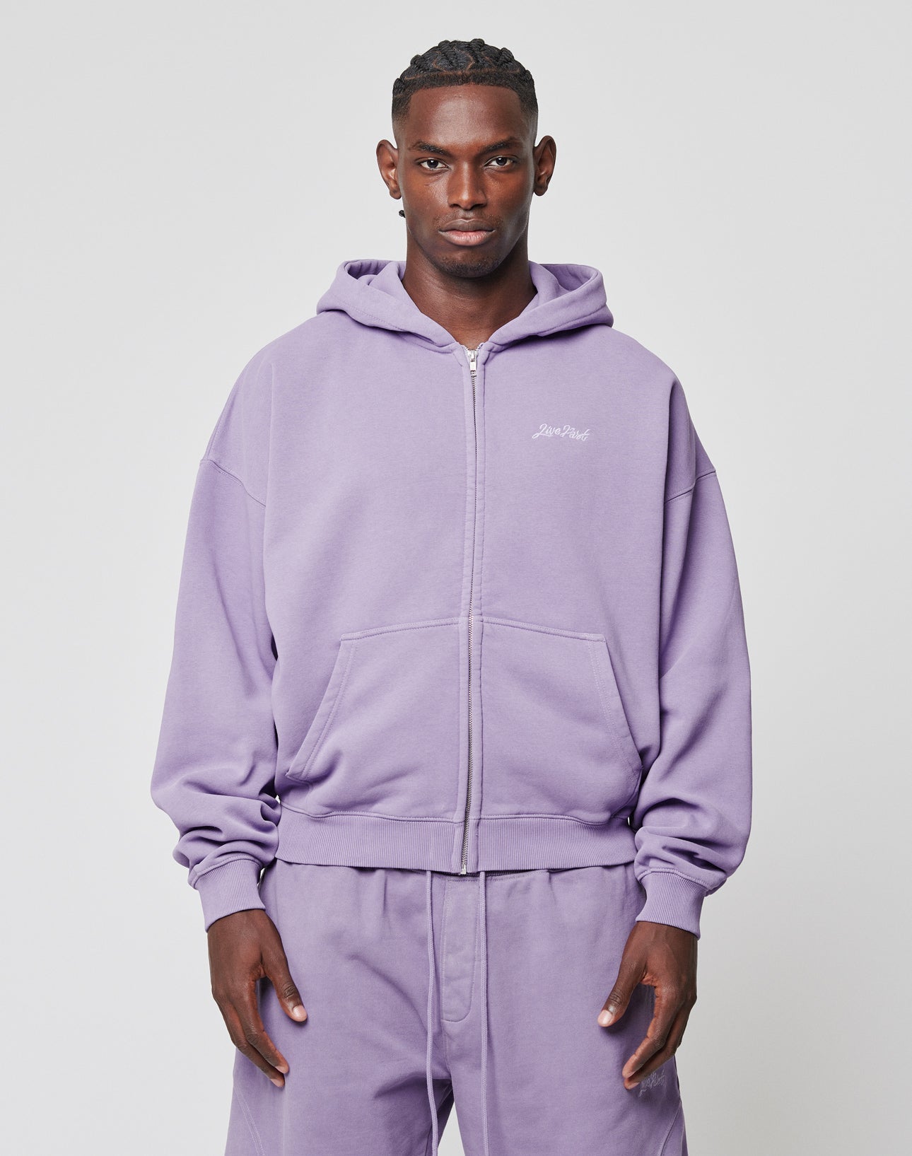 A person faces forward, wearing the LIVE FAST DIE YOUNG Basic Script Hooded Zip in light purple with Vintage-Waschung and matching sweatpants. The hoodie has a small white logo-stitch on the left chest. The background is plain and light-colored.