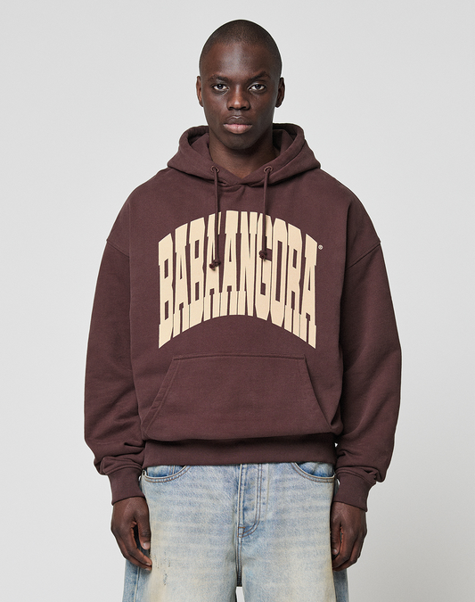 A person stands facing forward against a plain light gray background, wearing the LIVE FAST DIE YOUNG LFDY x Baba Angora Hoodie with large beige BABALANGORA letters on the chest, paired with light blue jeans. | model