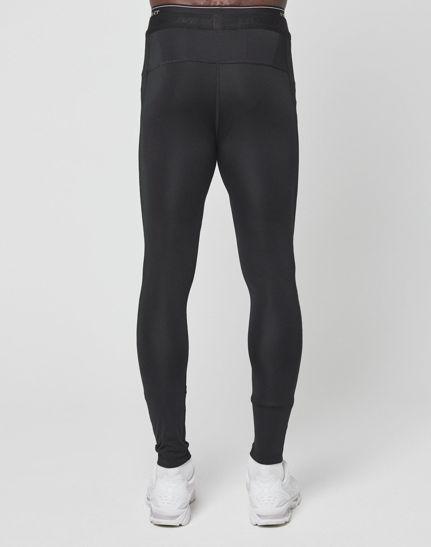 A person wearing LIVE FAST DIE YOUNGs Live Fast Performance Tights in slim fit black with an elastic waistband and white sneakers, shown from the back against a plain white background.
