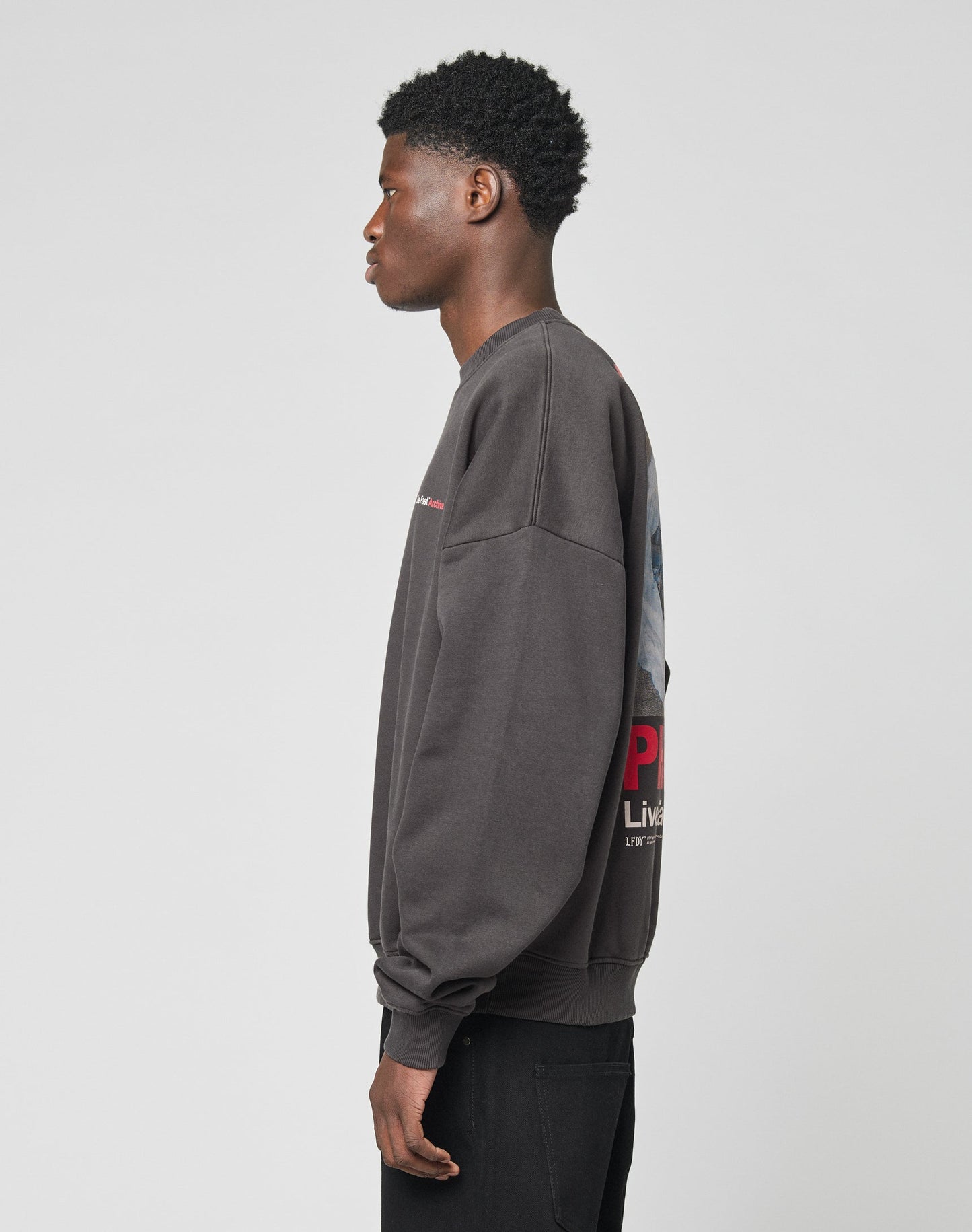 A young man stands in profile against a plain light background, wearing the LIVE FAST DIE YOUNG LFDY F*ck Sweater—an oversize fit dark gray Baumwolle sweatshirt with a graphic back design—and black pants.
