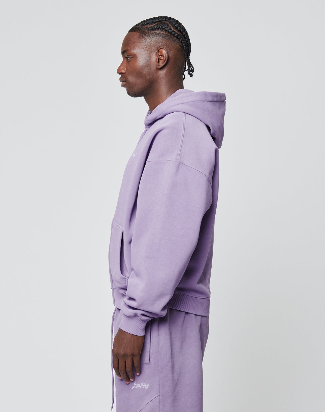 A person stands in profile against a plain background, wearing a lavender LIVE FAST DIE YOUNG Basic Script Hooded Zip hoodie with matching sweatpants. Their hair is styled in cornrows and their hands are by their sides.