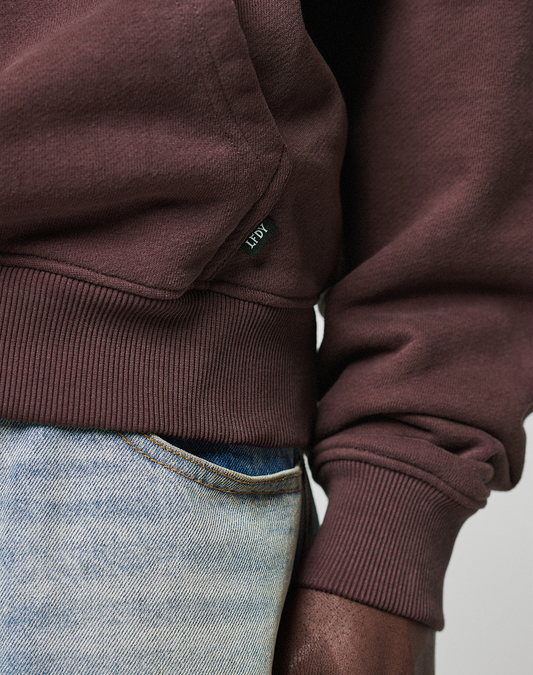 Close-up of a person wearing the maroon LFDY x Baba Angora Hoodie by LIVE FAST DIE YOUNG, featuring ribbed cuffs and a small logo tag, paired with light blue jeans. Only the hand and part of the torso are in view.